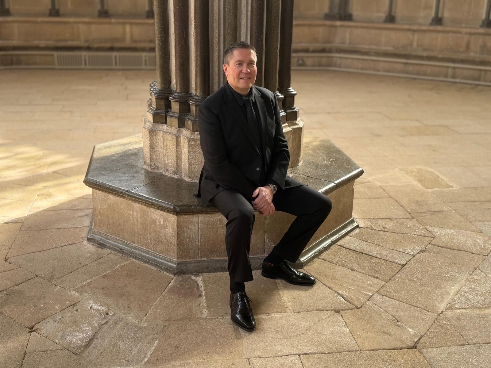 Matthew Minter seated in a stone cloister in a black suit.