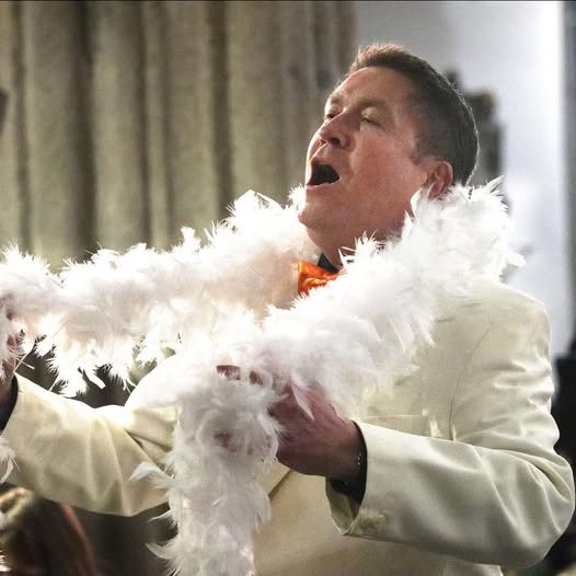 Matthew Minter singing in a white costume with a feather boa.