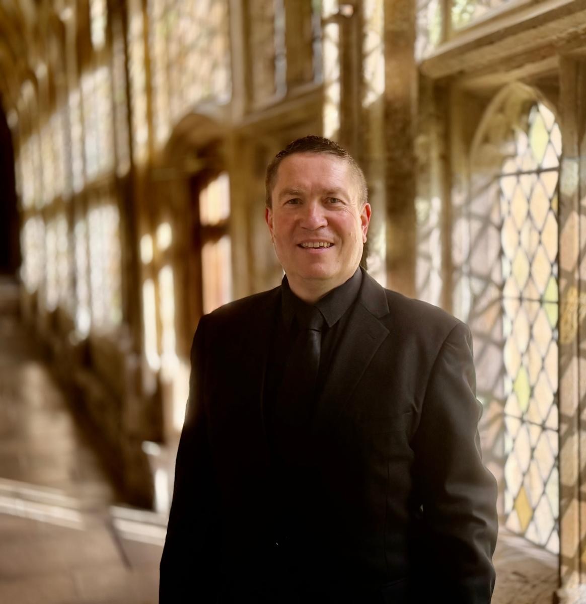 Matthew Minter standing in a softly lit stone corridor wearing a black suit.
